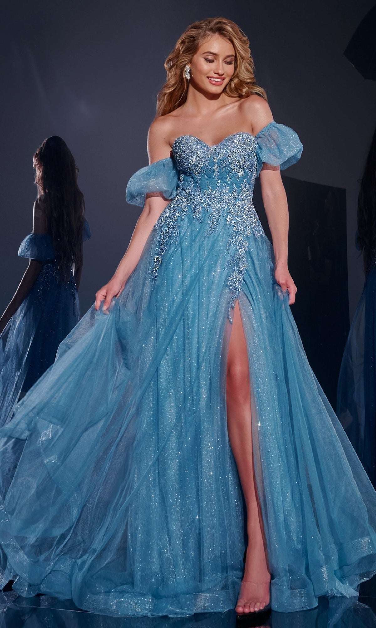 A woman poses elegantly on a dark reflective surface wearing the Jovani 40474 Long Blue Prom Dress with sparkling fabric, off-the-shoulder puff sleeves, and a high leg slit.